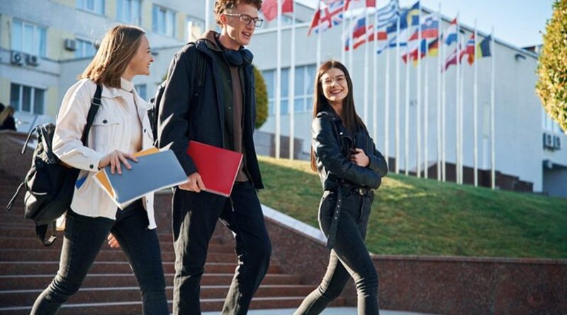 Trois étudiants Erasmus+ souriants marchant devant un bâtiment universitaire, avec des drapeaux européens en arrière-plan, symbolisant l’ouverture internationale de la France à travers deux millions de parcours Erasmus+