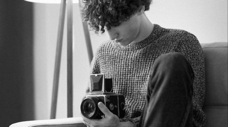 Homme assis sur un canapé, tenant un appareil photo argentique, en noir et blanc.