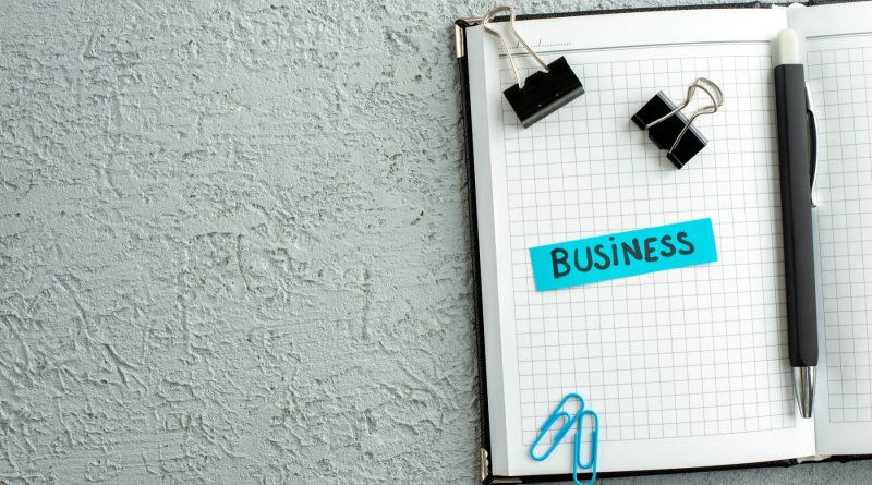 Carnet quadrillé ouvert avec un stylo, des trombones, des pinces noires et une étiquette bleue portant l’inscription « BUSINESS »