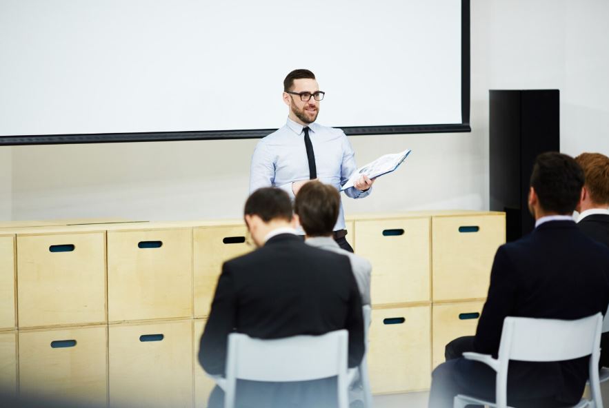 Un homme en tenue professionnelle, debout devant un écran de projection, tient des documents et s’adresse à un groupe de personnes assises