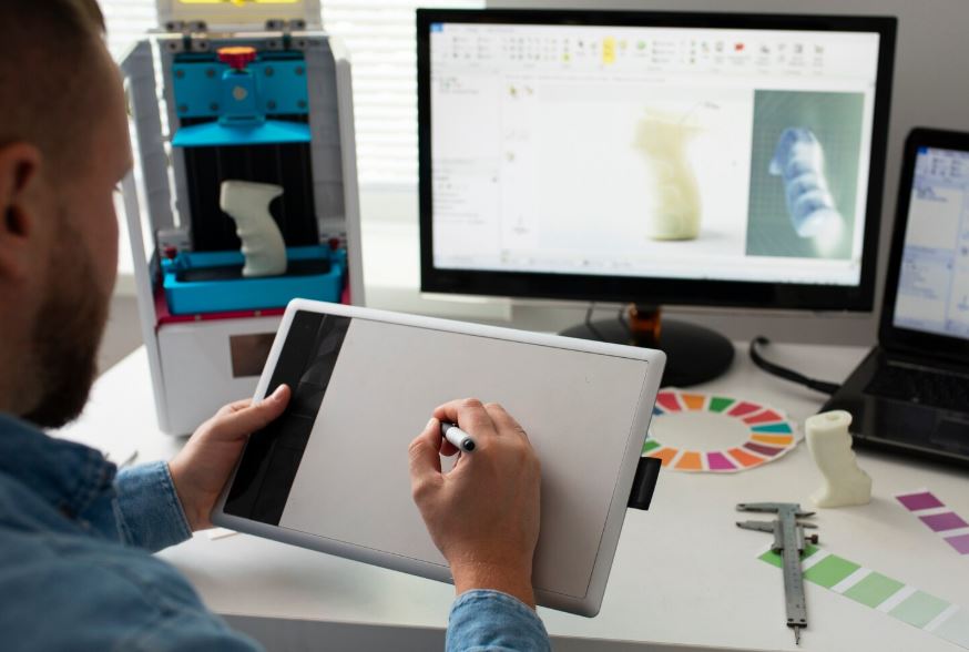 Designer utilisant une tablette graphique pour créer un modèle 3D affiché sur un ordinateur, dans un bureau équipé d’une imprimante 3D et d’outils de conception