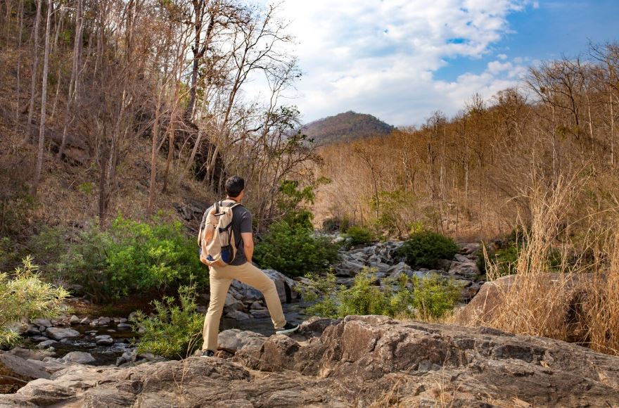 Randonneur avec sac à dos observant un paysage naturel composé de rochers, d’un ruisseau et d’une forêt sèche, sous un ciel partiellement nuageux