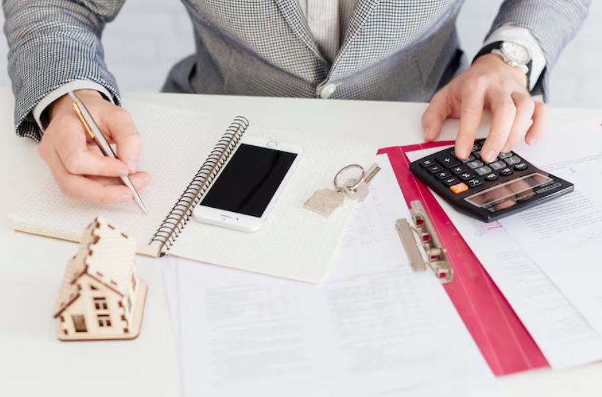 Un investisseur immobilier utilise une calculatrice et prend des notes dans un carnet à côté d'une maquette de maison