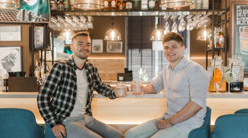 Deux jeunes hommes souriants sont assis au comptoir d'un bar tenant chacun un verre à la main