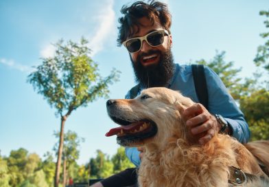 Un homme barbu souriant avec des lunettes de soleil caresse son chien Golden Retriever en extérieur sous un ciel bleu