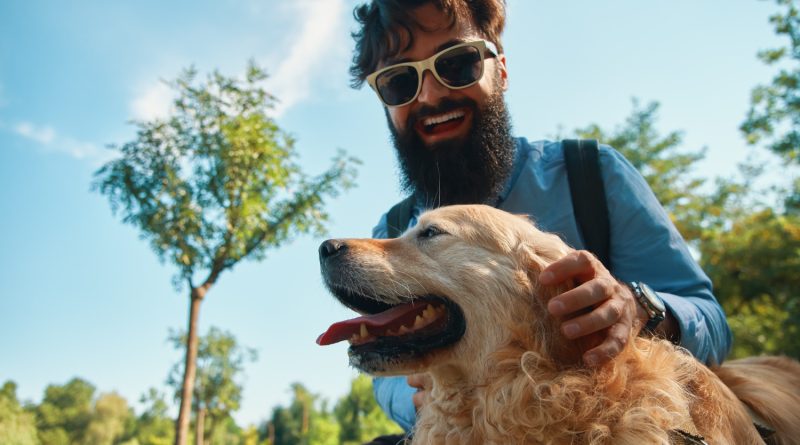 Un homme barbu souriant avec des lunettes de soleil caresse son chien Golden Retriever en extérieur sous un ciel bleu