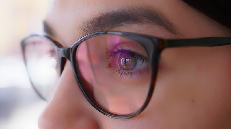 œil d'une femme portant des lunettes de vue à monture noire avec des reflets roses et violets sur les verres