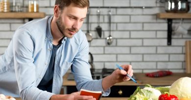 Un homme dans une cuisine note soigneusement ses courses dans un carnet tout en examinant des légumes frais posés sur le plan de travail