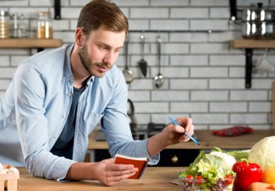 Un homme dans une cuisine note soigneusement ses courses dans un carnet tout en examinant des légumes frais posés sur le plan de travail