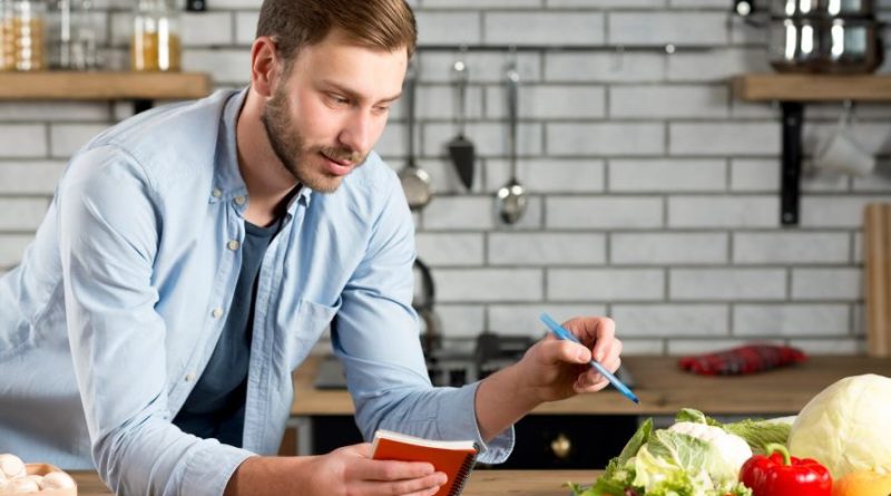 Un homme dans une cuisine note soigneusement ses courses dans un carnet tout en examinant des légumes frais posés sur le plan de travail