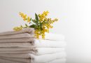 Une pile de serviettes de bain blanches et propres, soigneusement pliées sur un fond neutre et épuré. Un petit bouquet de fleurs sauvages jaunes et vertes est délicatement posé sur le dessus de la pile