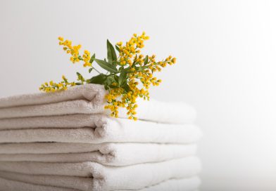 La Berge : comment le linge de bain haut de gamme transforme l’expérience de la salle de bain Une pile de serviettes de bain blanches et propres, soigneusement pliées sur un fond neutre et épuré. Un petit bouquet de fleurs sauvages jaunes et vertes est délicatement posé sur le dessus de la pile
