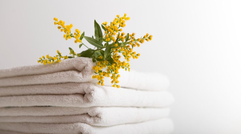 Une pile de serviettes de bain blanches et propres, soigneusement pliées sur un fond neutre et épuré. Un petit bouquet de fleurs sauvages jaunes et vertes est délicatement posé sur le dessus de la pile