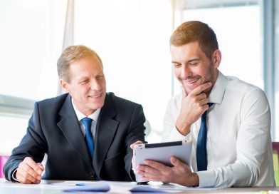La gestion de patrimoine à l’heure de la démocratisation de l’investissement Deux conseillers financiers en costume sourient en regardant ensemble une tablette numérique dans un bureau lumineux.