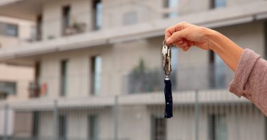 Une main tendue tient un trousseau de clés avec un pompon bleu devant un immeuble résidentiel moderne et flou en arrière-plan