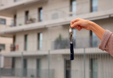 Une main tendue tient un trousseau de clés avec un pompon bleu devant un immeuble résidentiel moderne et flou en arrière-plan