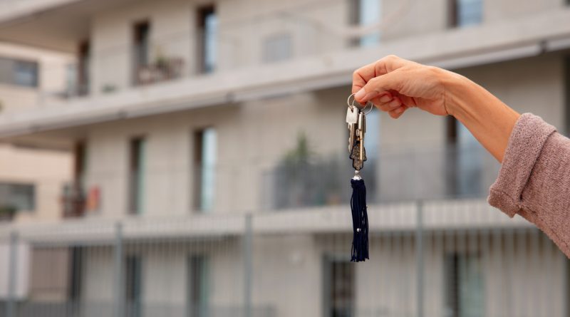 Une main tendue tient un trousseau de clés avec un pompon bleu devant un immeuble résidentiel moderne et flou en arrière-plan
