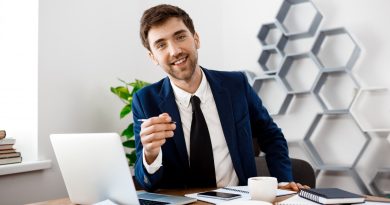 un jeune homme souriant à son bureau, habillé de manière professionnelle dans un costume bleu marine, une chemise blanche et une cravate noire. Il tient un stylo et regarde directement l'objectif