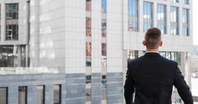 un homme d'affaires vu de dos, vêtu d'un costume sombre, contemplant des immeubles modernes depuis un balcon