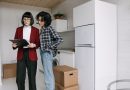 une agente immobilière en veste rouge examinant des documents sur un porte-bloc avec une cliente dans une cuisine moderne en plein déménagement.