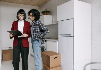 Pourquoi la gestion locative devient un levier stratégique pour valoriser son patrimoine immobilier une agente immobilière en veste rouge examinant des documents sur un porte-bloc avec une cliente dans une cuisine moderne en plein déménagement.