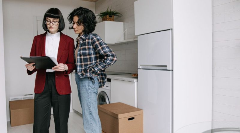 Pourquoi la gestion locative devient un levier stratégique pour valoriser son patrimoine immobilier une agente immobilière en veste rouge examinant des documents sur un porte-bloc avec une cliente dans une cuisine moderne en plein déménagement.