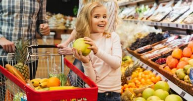 Une petite fille souriante tenant un fruit dans un rayon de supermarché aux côtés de ses parents.