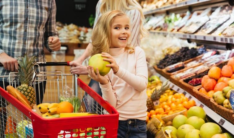Une petite fille souriante tenant un fruit dans un rayon de supermarché aux côtés de ses parents.