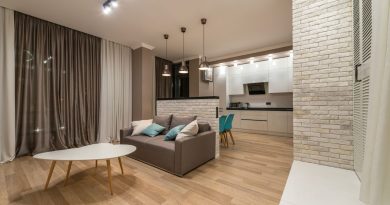 un salon avec un canapé gris et une table basse en bois, ouvert sur une cuisine équipée séparée par une muret en briques blanches