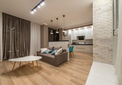 un salon avec un canapé gris et une table basse en bois, ouvert sur une cuisine équipée séparée par une muret en briques blanches