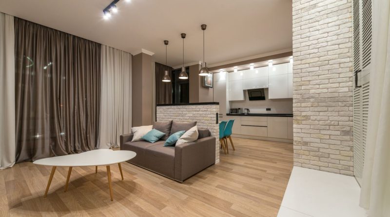 un salon avec un canapé gris et une table basse en bois, ouvert sur une cuisine équipée séparée par une muret en briques blanches