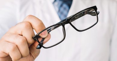 Un opticien en blouse blanche tend une paire de lunettes de vue à monture noire rectangulaire vers l'avant.