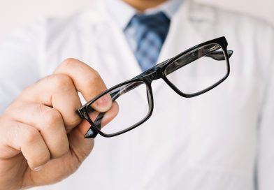 Un opticien en blouse blanche tend une paire de lunettes de vue à monture noire rectangulaire vers l'avant.