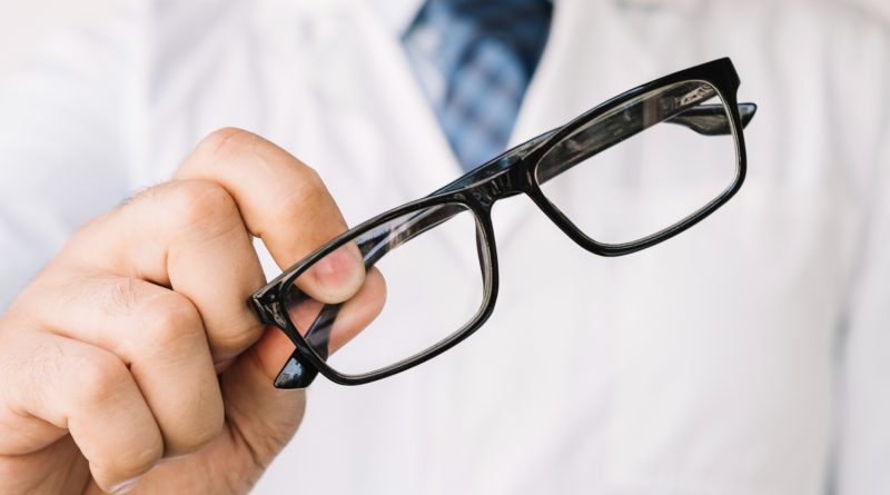 Un opticien en blouse blanche tend une paire de lunettes de vue à monture noire rectangulaire vers l'avant.