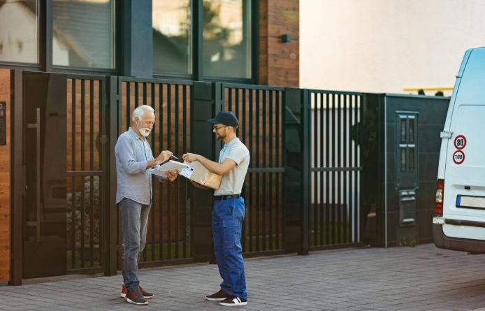 Un livreur remet un colis à un client âgé qui signe le bon de livraison devant sa maison, près d'une camionnette blanche stationnée dans la rue