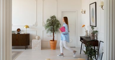 Une femme portant une chemise bleue et tenant un classeur rose se déplace à travers une vaste pièce élégante de style haussmannien, aux murs blancs ornés de moulures classiques.