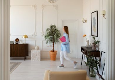 Cosy Home : la location meublée à Paris, une solution pour accompagner la mobilité professionnelle Une femme portant une chemise bleue et tenant un classeur rose se déplace à travers une vaste pièce élégante de style haussmannien, aux murs blancs ornés de moulures classiques.