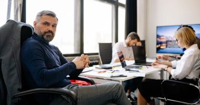 Un cadre aux cheveux et à la barbe poivre et sel siège dans un bureau moderne, les mains croisées, tandis que ses collaborateurs travaillent en arrière-plan.
