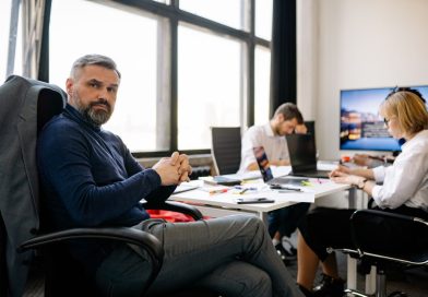 Un cadre aux cheveux et à la barbe poivre et sel siège dans un bureau moderne, les mains croisées, tandis que ses collaborateurs travaillent en arrière-plan.