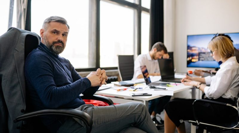 Un cadre aux cheveux et à la barbe poivre et sel siège dans un bureau moderne, les mains croisées, tandis que ses collaborateurs travaillent en arrière-plan.