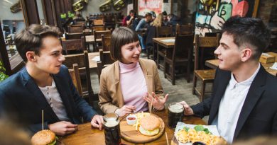 Un groupe de jeunes amis discute joyeusement autour d'une table en bois dans un bar-restaurant chaleureux, partageant des burgers, des frites et des chopes de bière.