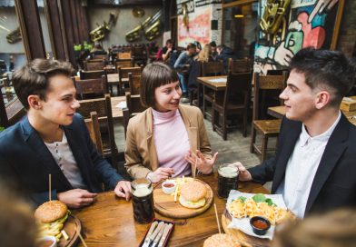 Thierry Di Litta et Barabaar : trois quartiers de Lyon, trois façons de vivre la convivialité Un groupe de jeunes amis discute joyeusement autour d'une table en bois dans un bar-restaurant chaleureux, partageant des burgers, des frites et des chopes de bière.