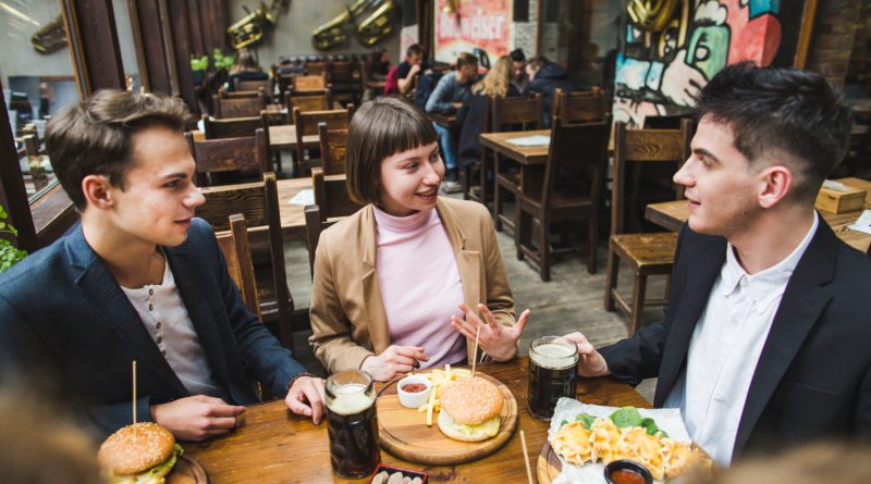 Un groupe de jeunes amis discute joyeusement autour d'une table en bois dans un bar-restaurant chaleureux, partageant des burgers, des frites et des chopes de bière.